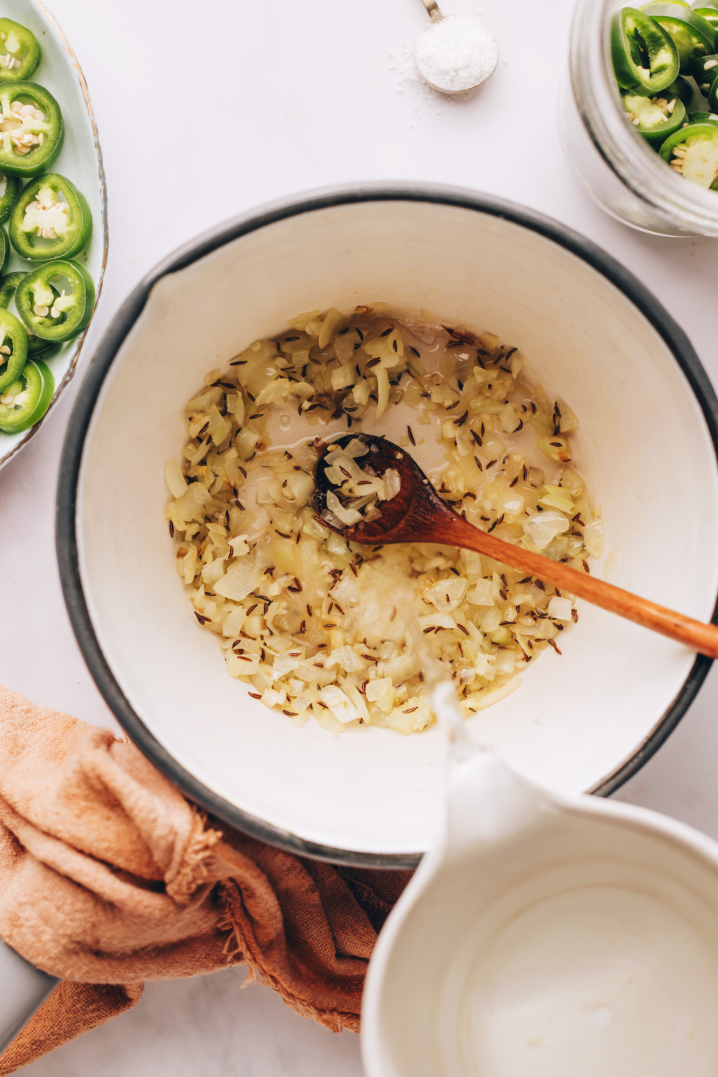 Pouring water into a saucepan of sautéed onion, garlic, and cumin seeds