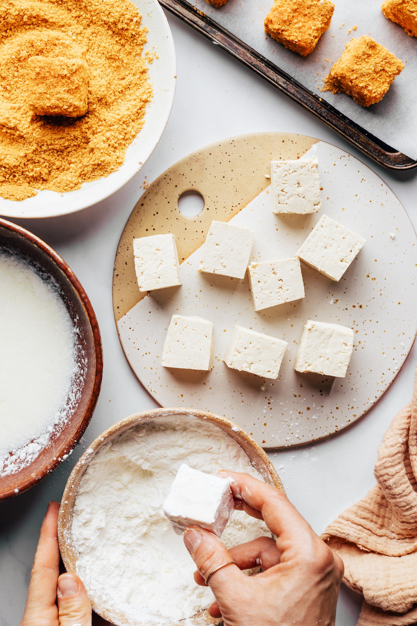 Dipping cubed tofu into cornstarch next to bowls of vegan "buttermilk" and a cashew coating