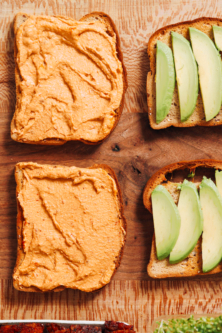 Four slices of bread — two topped with avocado and tomato and the other two with hummus, cucumber, tempeh bacon, and alfalfa sprouts