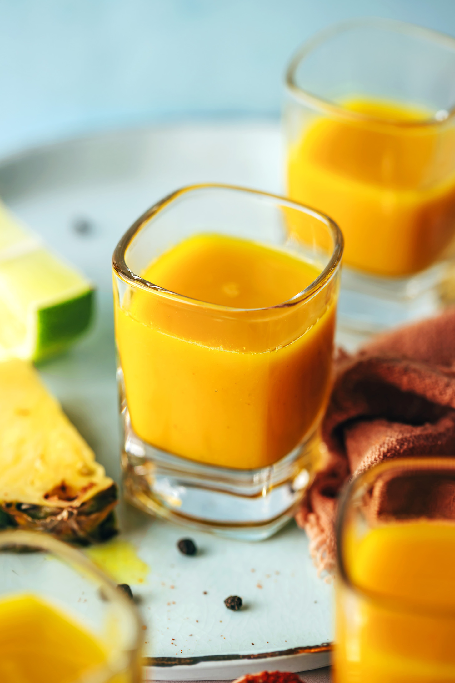 Shot glasses of our easy fire cider recipe on a plate next to black peppercorns, pineapple, and lime juice