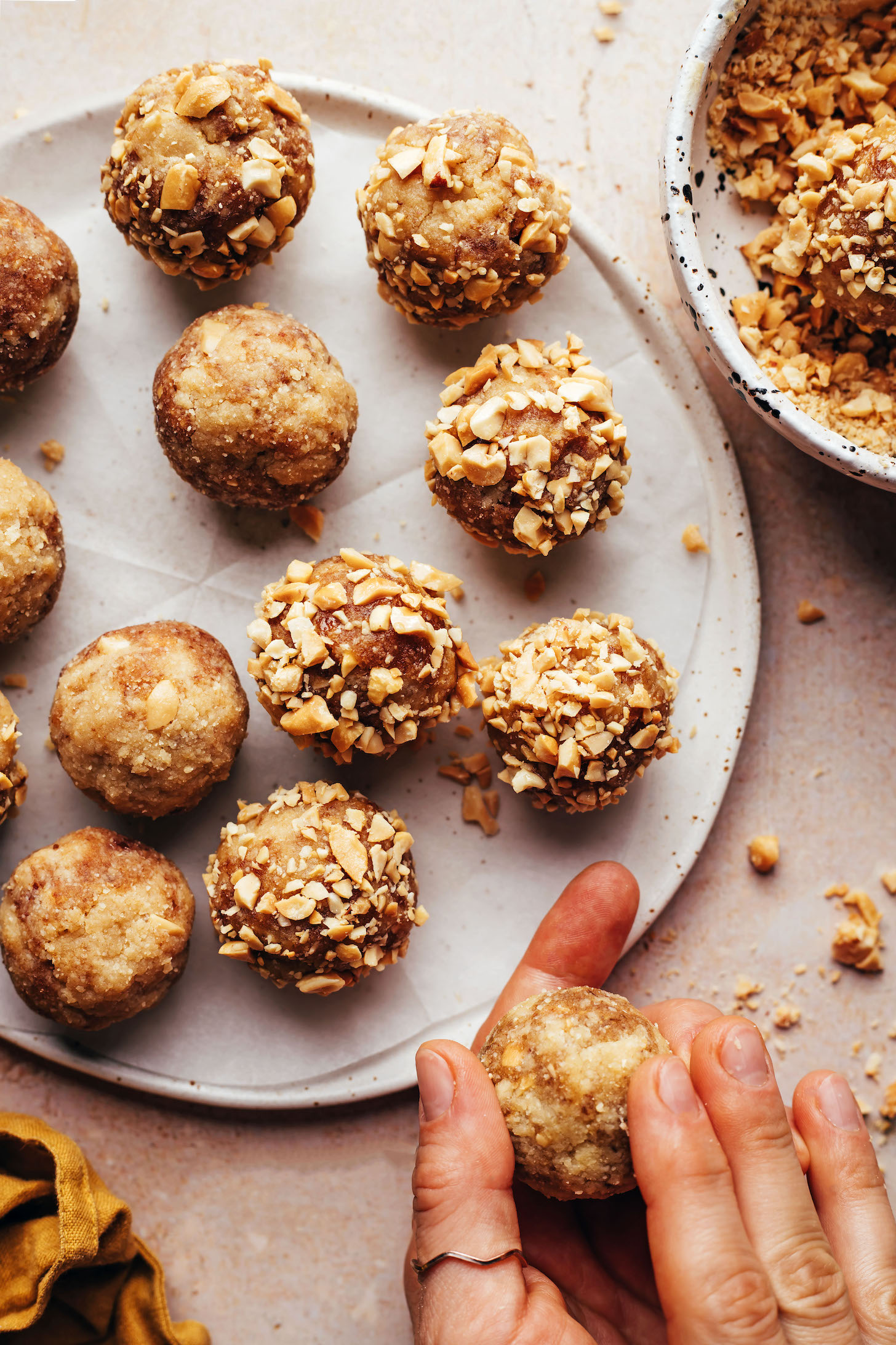 Rolling a snickers energy bite next to a plate of energy bites and bowl of chopped peanuts