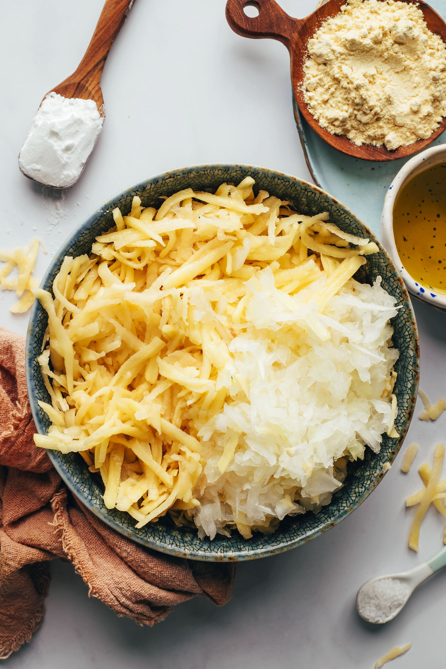 Bowl of shredded onions and potatoes surrounded by potato starch, oil, salt, and chickpea flour