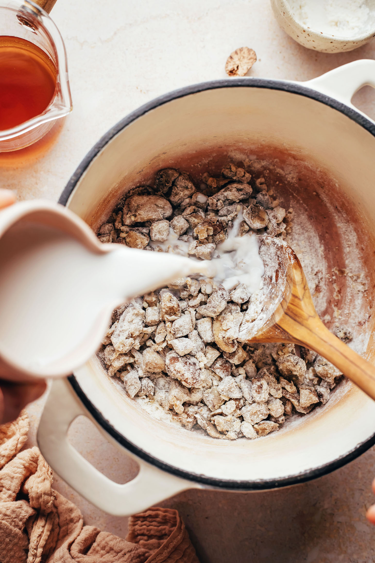 Pouring dairy-free milk into a pot of sautéed mushrooms coated in gluten-free flour