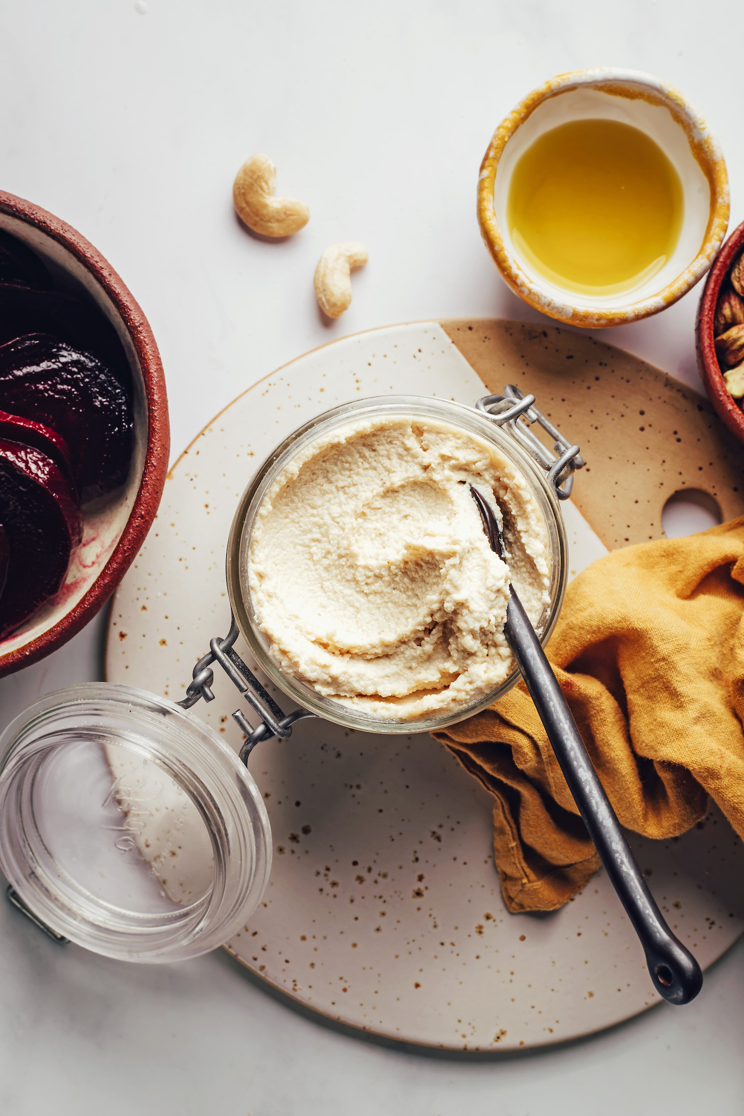 Overhead shot of bowls of olive oil, cashews, and roasted beets, plus a spoon in a jar of homemade vegan goat cheese