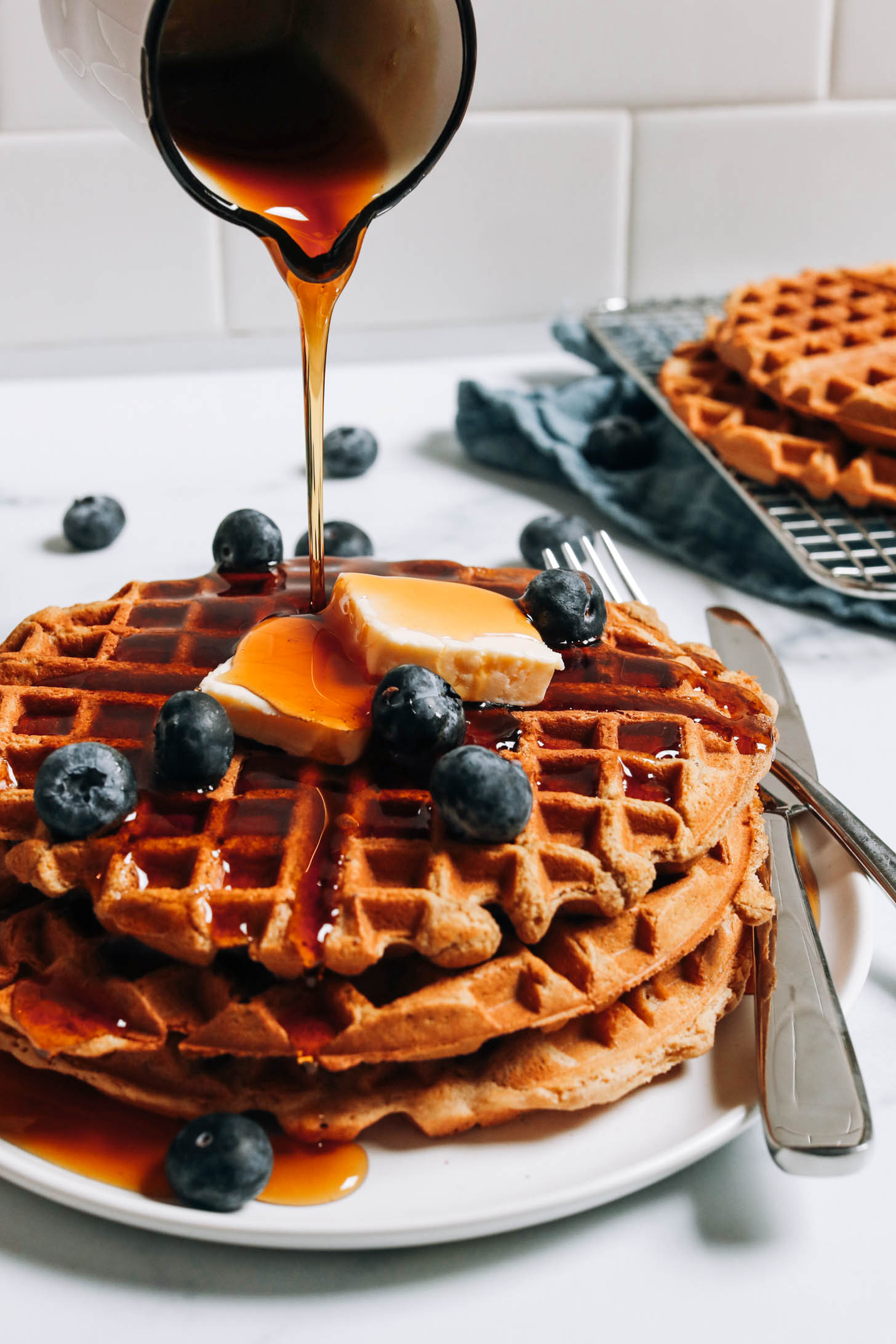 Drizzling maple syrup onto a stack of waffles topped with vegan butter and blueberries