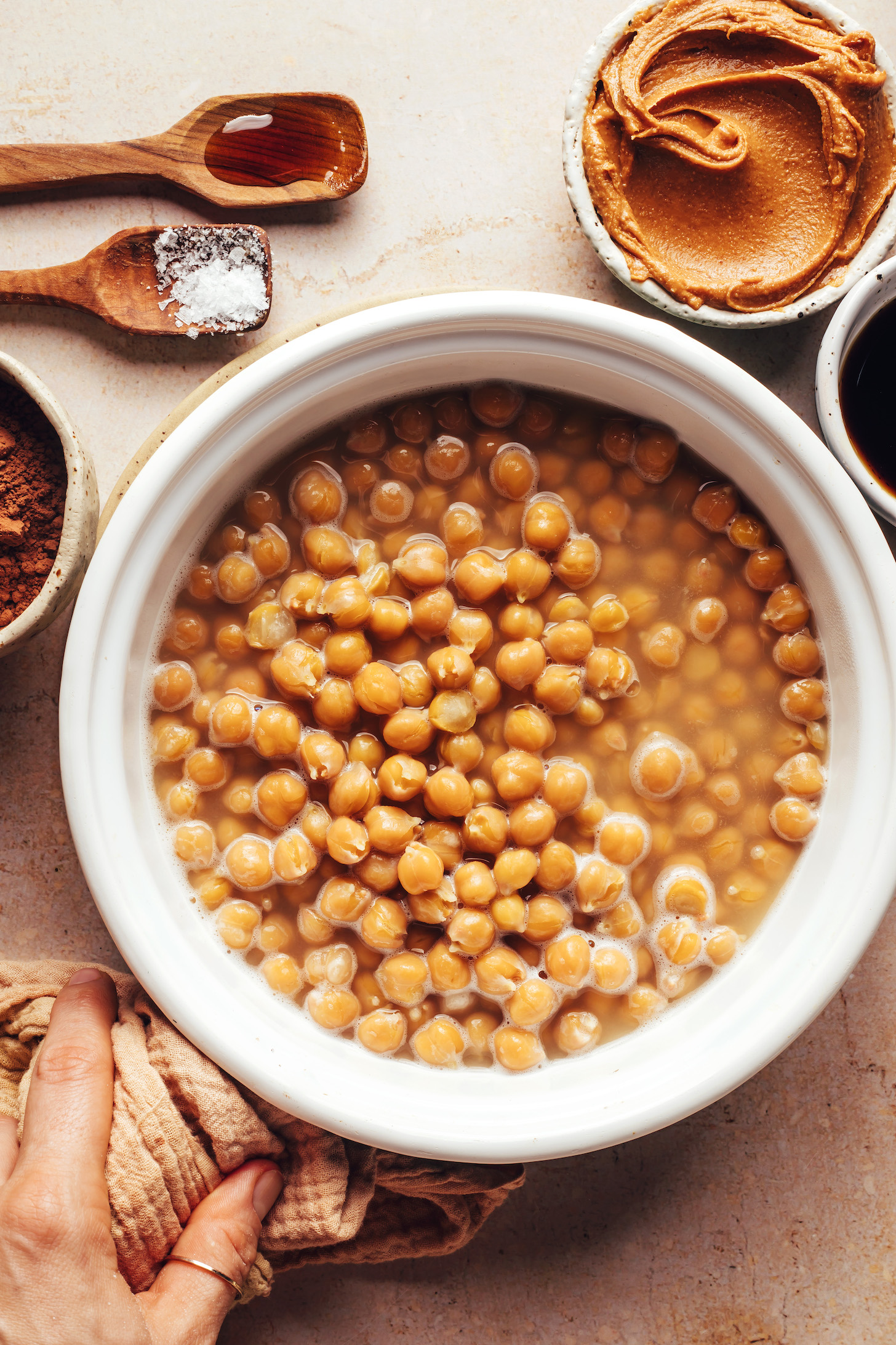 Saucepan of chickpeas surrounded by spoons and bowls of vanilla, salt, cocoa powder, nut butter, and vanilla