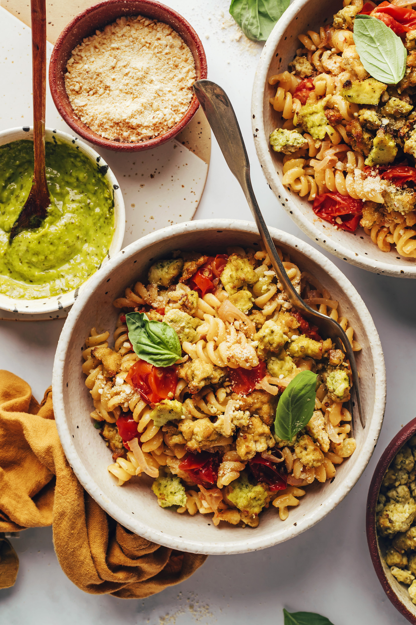 Bowls of our easy pesto pasta with crispy tofu and veggies next to more pesto and vegan parmesan