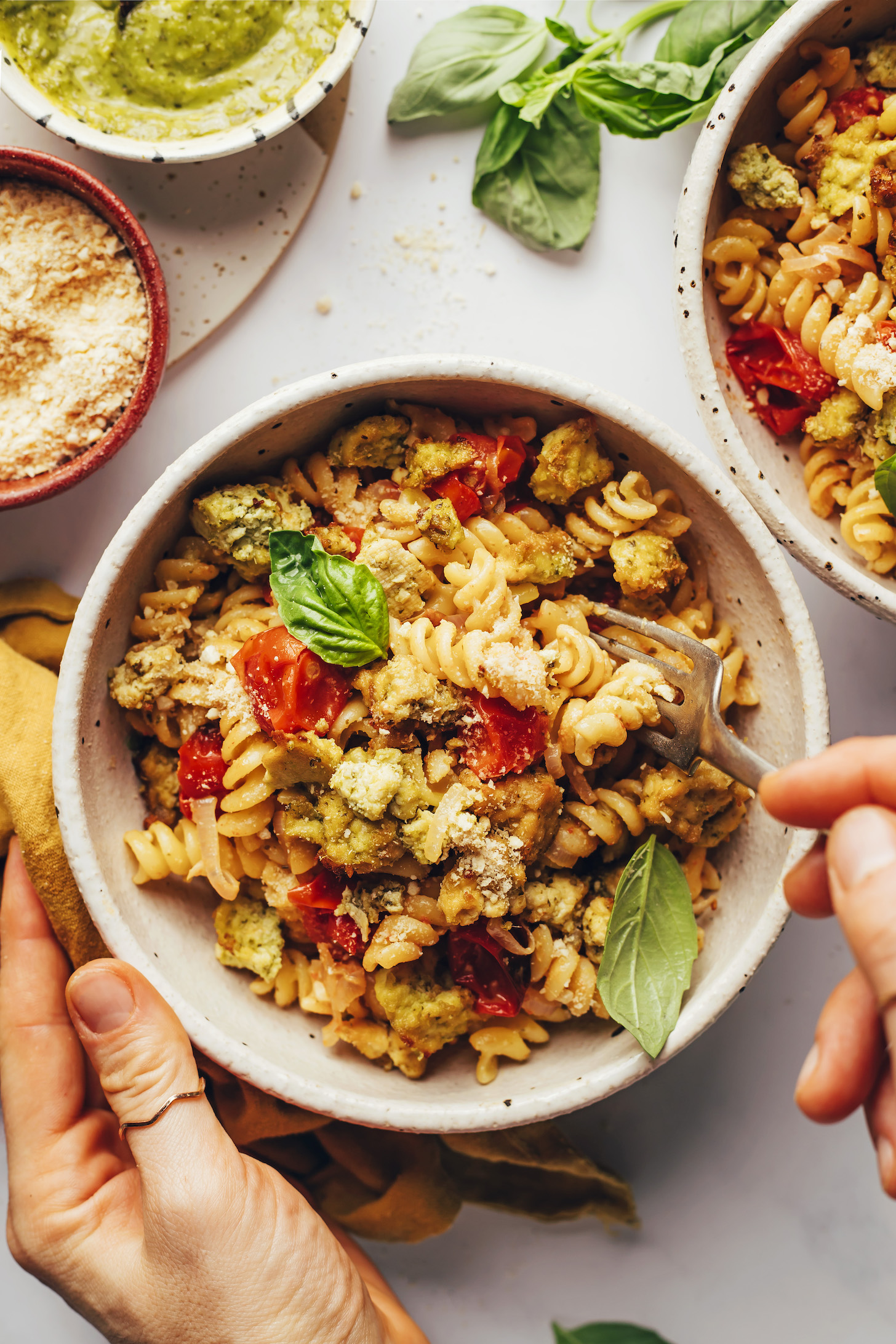 Hands holding the side of a bowl and a fork picking up a bite of pesto pasta with crispy tofu