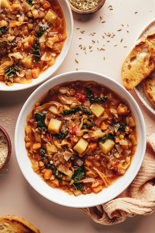 Two bowls of cabbage, fennel, and lentil soup next to a plate of sliced bread
