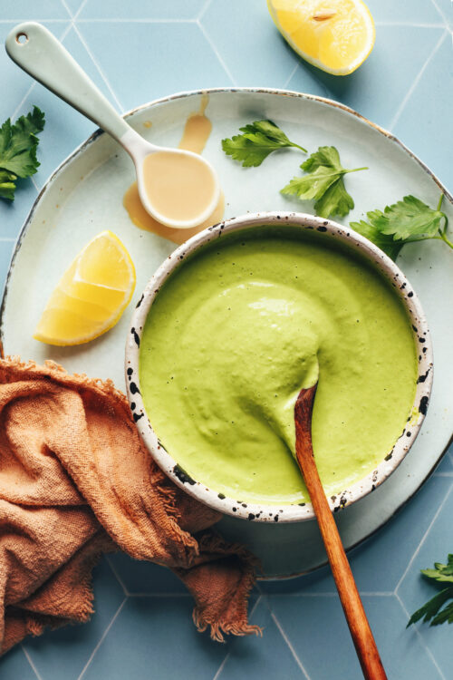 Bowl of herby green tahini sauce next to parsley, lemon, and tahini