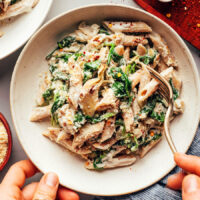 Hand holding a fork in a bowl of vegan spinach artichoke pasta