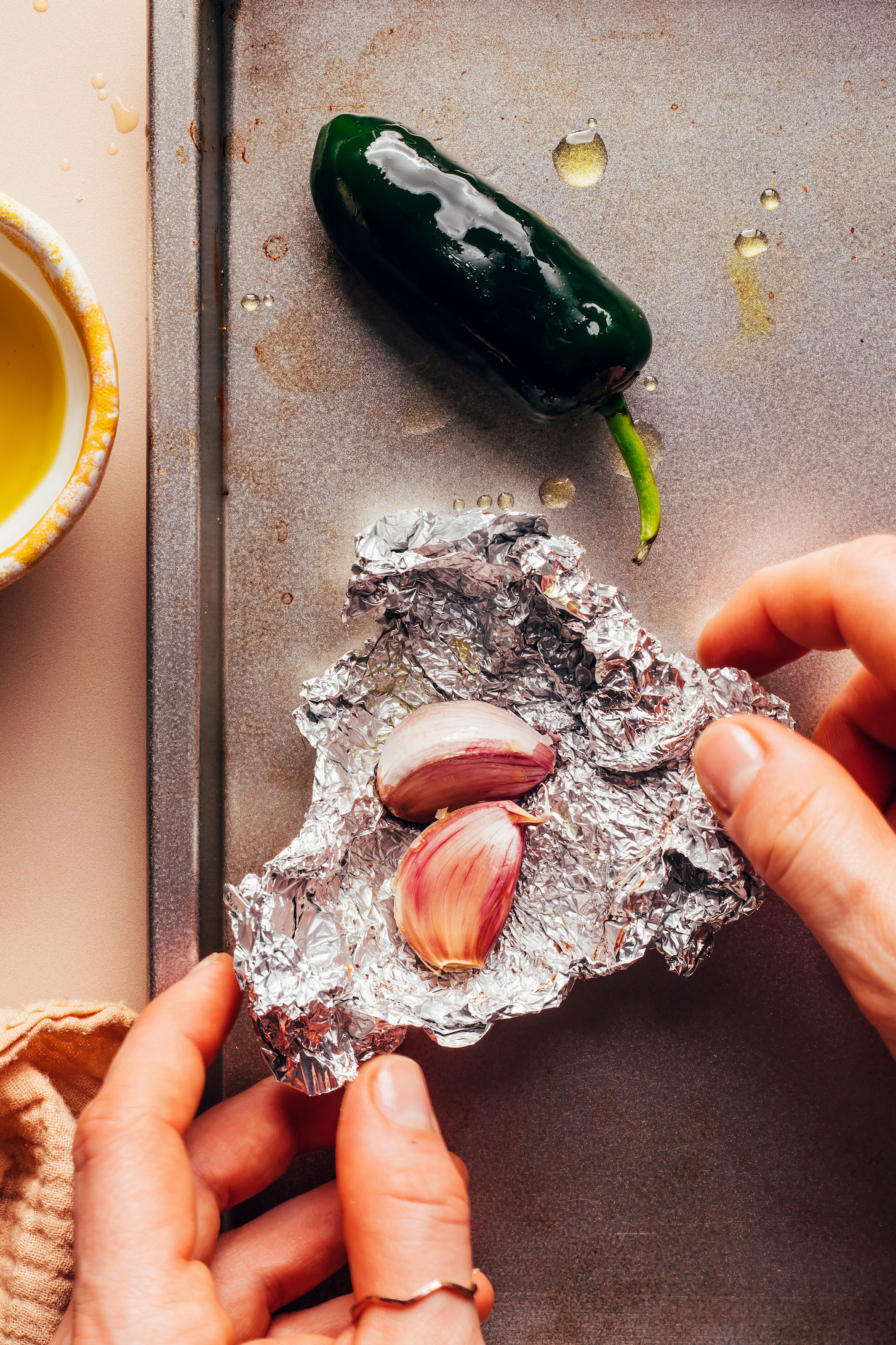 Jalapeño on a baking sheet next to hands wrapping whole garlic cloves in foil
