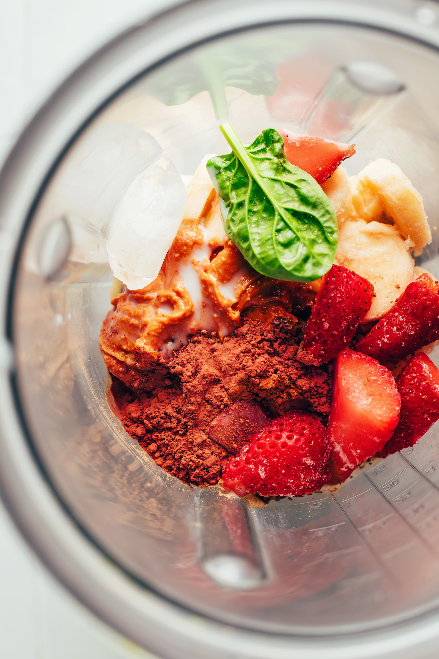 Blender with frozen strawberries, frozen banana, cacao powder, nut butter, spinach, and ice