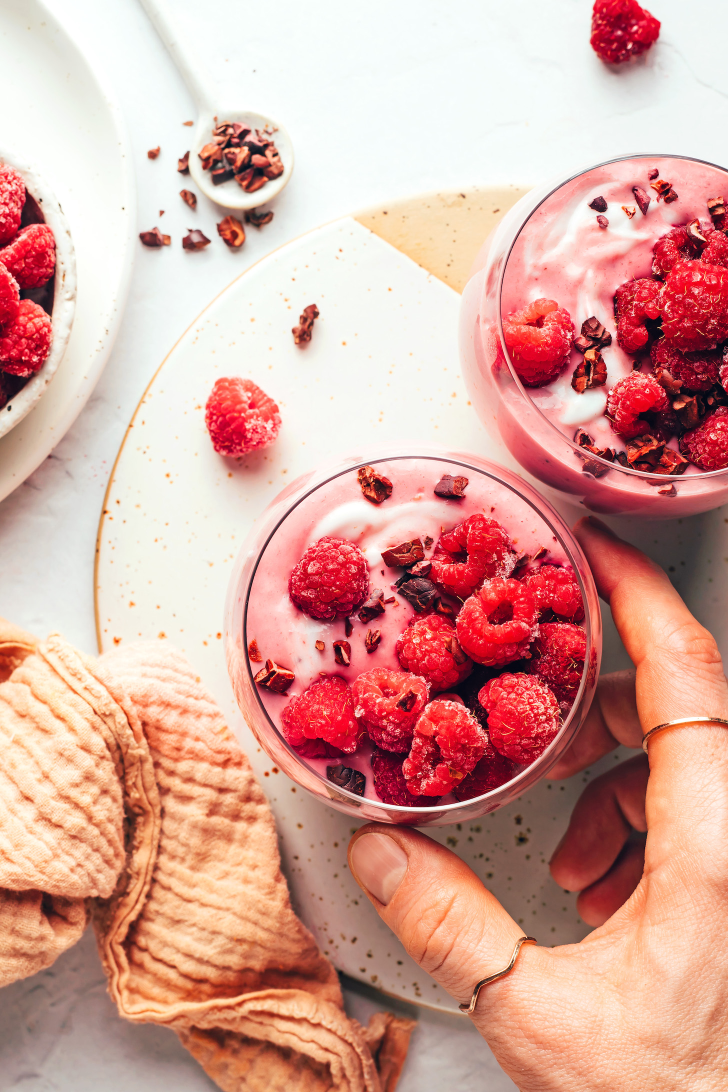 Hand reaching in to pick up a glass of raspberry banana smoothie topped with raspberries and cacao nibs
