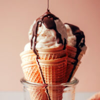 Drizzling chocolate magic shell onto scoops of no-churn vegan vanilla ice cream resting in cones