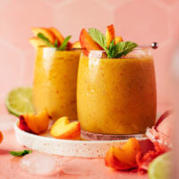 Two glasses of our frozen peach daiquiri with peach slices, mint, and limes