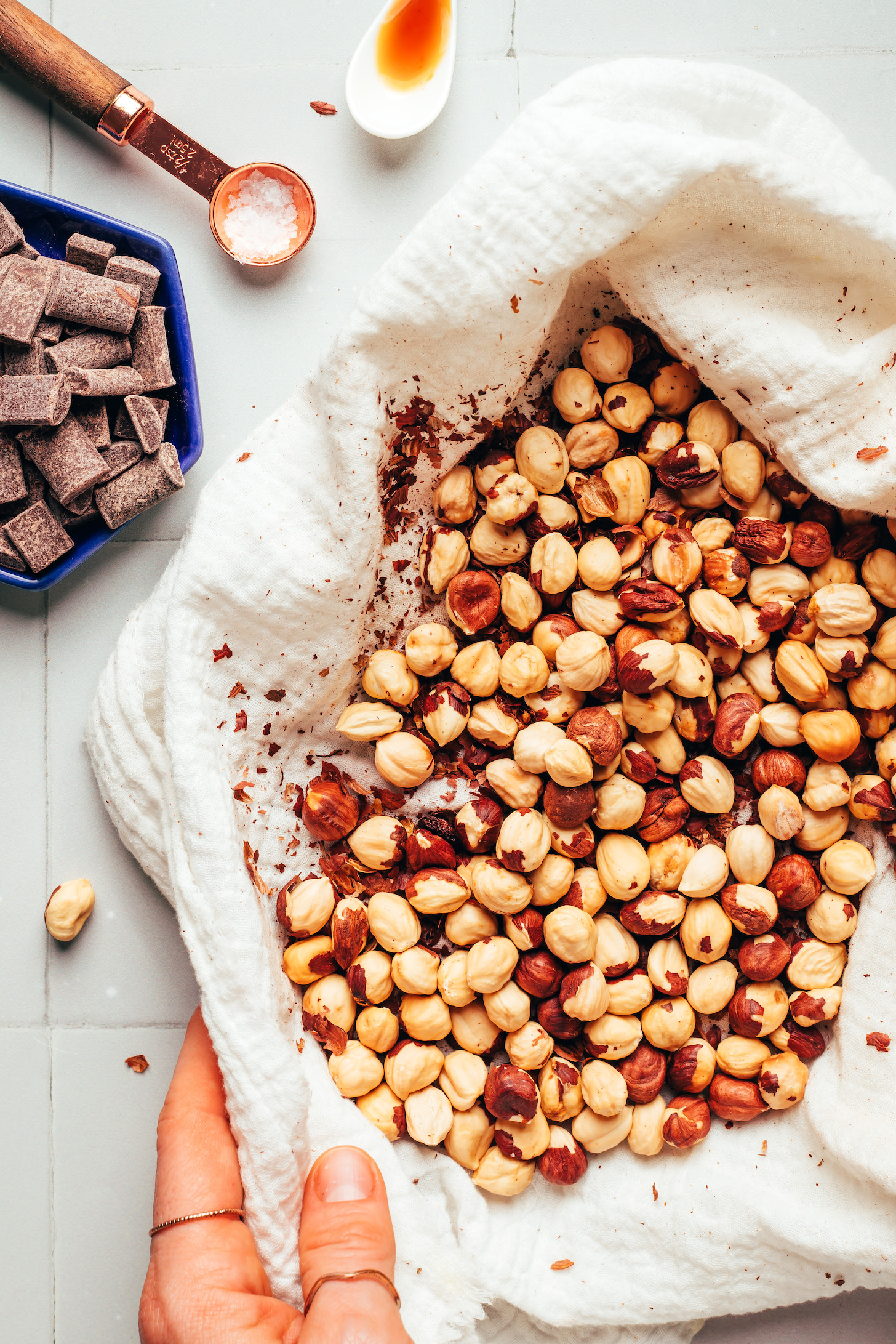 Chocolate chunks, salt, and vanilla next to a batch of roasted hazelnuts in a clean dish towel used to remove the skins