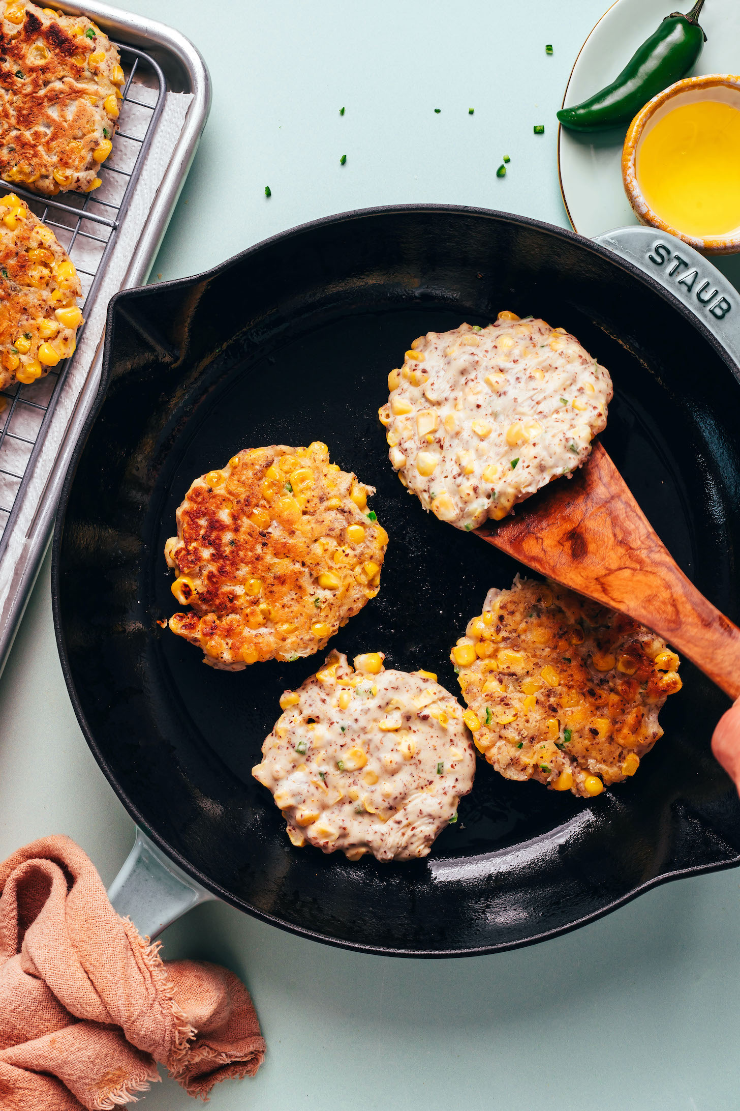 Using a wooden spatula to flip a vegan jalapeño corn fritter in a cast iron skillet with more fritters cooking